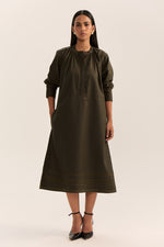 Load image into Gallery viewer, A woman modeling a dark olive green midi dress with three-quarter sleeves, a gathered neckline, and subtle horizontal striped detailing near the hem, paired with black ankle-strap heels against a plain light beige studio background.