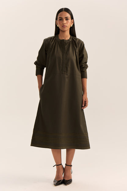 A woman modeling a dark olive green midi dress with three-quarter sleeves, a gathered neckline, and subtle horizontal striped detailing near the hem, paired with black ankle-strap heels against a plain light beige studio background.