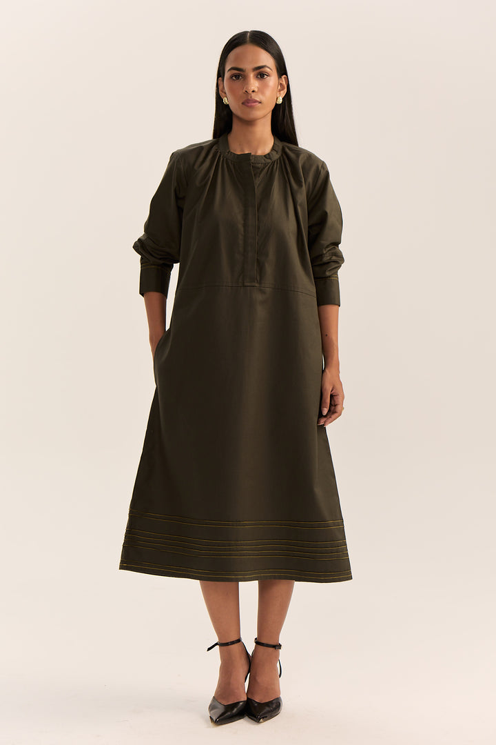 A woman modeling a dark olive green midi dress with three-quarter sleeves, a gathered neckline, and subtle horizontal striped detailing near the hem, paired with black ankle-strap heels against a plain light beige studio background.