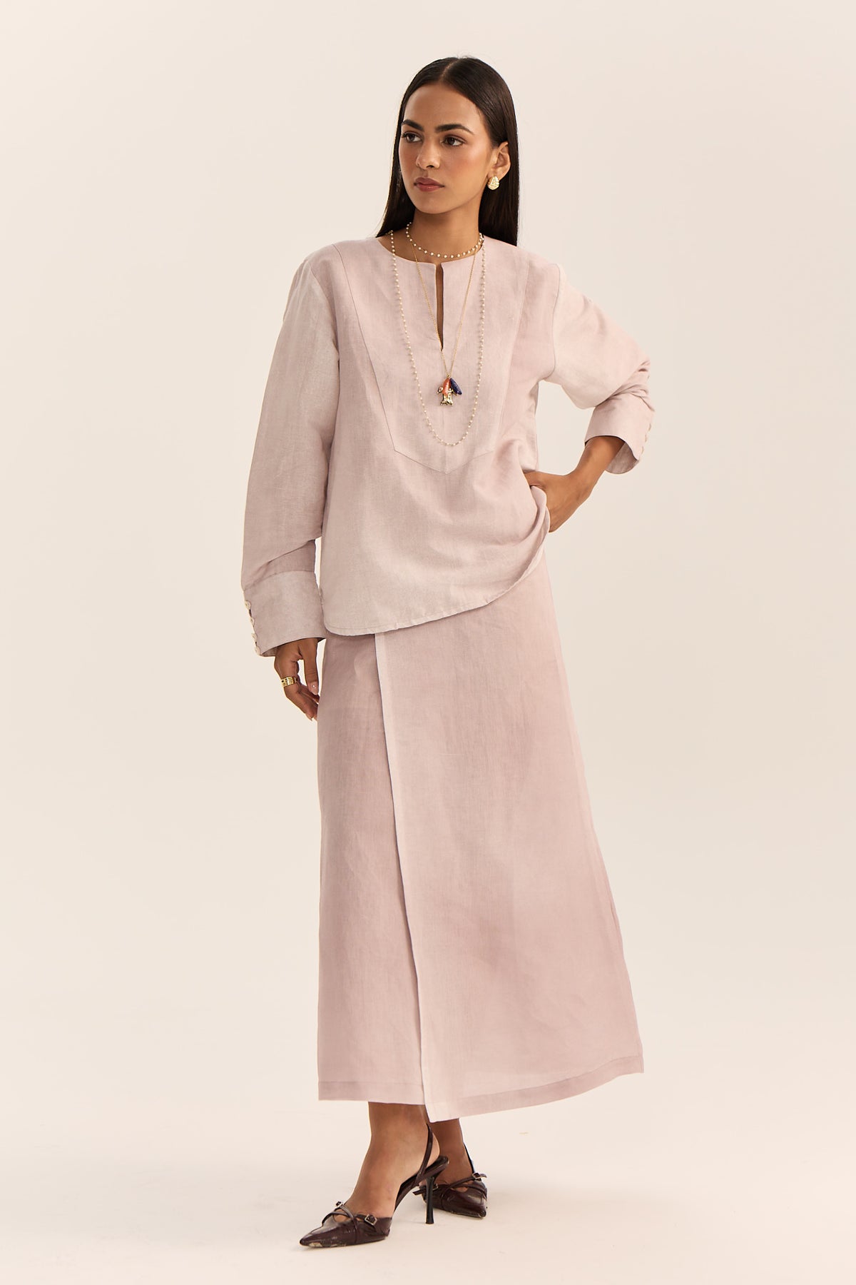 A woman modeling a light pink two-piece outfit consisting of a long-sleeve tunic top with a split neckline and a matching wide-leg skirt, photographed against a solid light beige studio background.