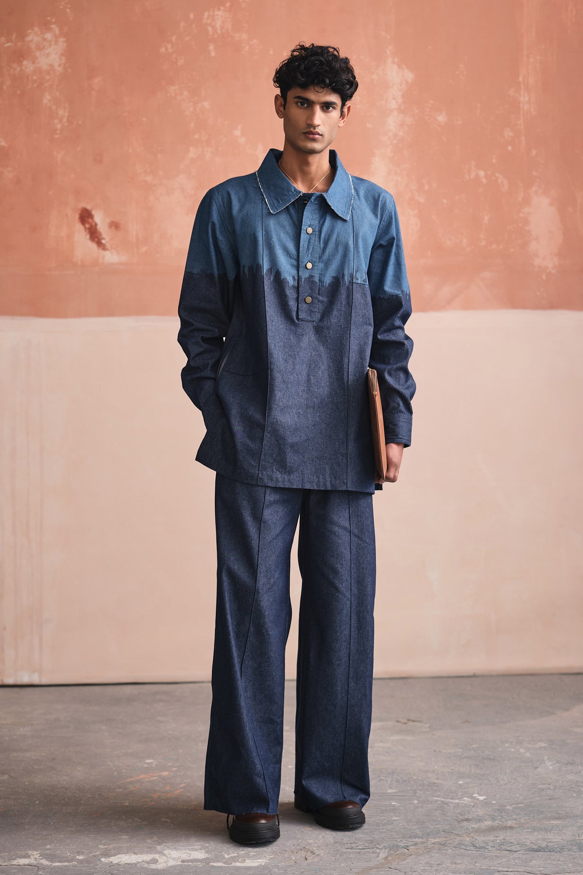 A person wearing a blue denim kurta set with a collar neckline, long sleeves, and matching pants standing against a pink wall.