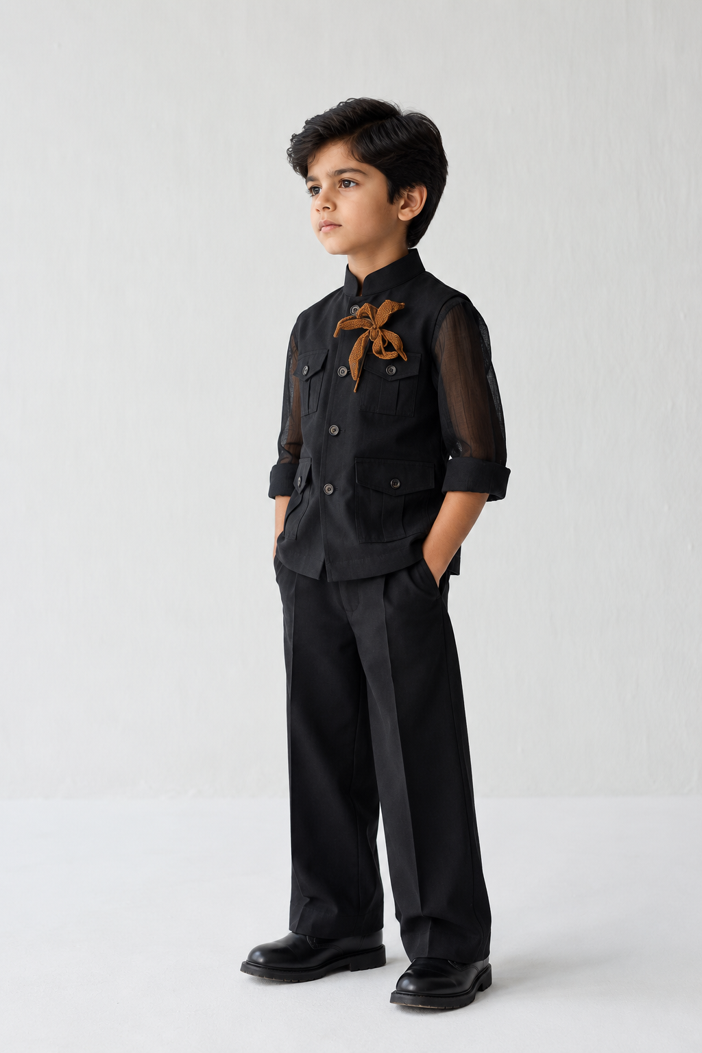 A young boy modeling a black traditional Indian outfit consisting of a long-sleeve button-up jacket with sheer sleeves and a brown embroidered motif on the chest, paired with black trousers and black shoes, against a plain light gray background.