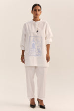 Load image into Gallery viewer, A model stands against a plain light beige studio background wearing a white kurta-style top with a blue architectural graphic print on the front, paired with white pants and black pointed heels.