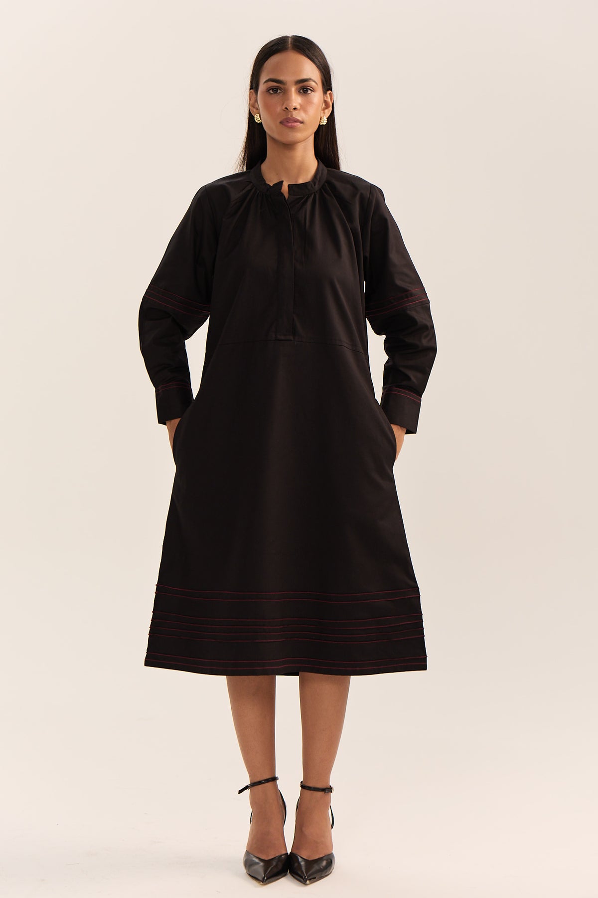 A woman modeling a black long-sleeve tunic dress with subtle red embroidered stripes near the hem and cuffs, paired with black high heels, against a plain light beige studio background.