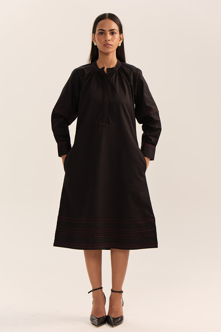 A woman modeling a black long-sleeve tunic dress with subtle red embroidered stripes near the hem and cuffs, paired with black high heels, against a plain light beige studio background.